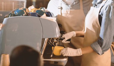 Barista yeni normal ve covid-19 salgını sırasında çalışıyor ve kahve yapıyor. Önlüklü, eldivenli ve koruyucu maskeli genç Afrikalı Amerikalı kadın ve adam kafede espresso makinesi kullanıyor.