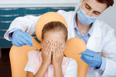 Dişçi ziyareti, bakıcılık ve korku. Korkmuş küçük çocuk stomatoloji sandalyesinde gözlerini kapar, beyaz önlüklü yakışıklı doktor, koruyucu maske ve lastik eldivenler tedaviye başlar, kopyalama alanı
