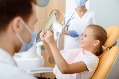 Harika bir iş, sağlıklı mükemmel dişler ve profesyonel doktor. Koruyucu maskeli genç bir dişçi, kliniğin iç kısımlarında elinde ayna olan gülümseyen küçük bir hastaya bakıyor.