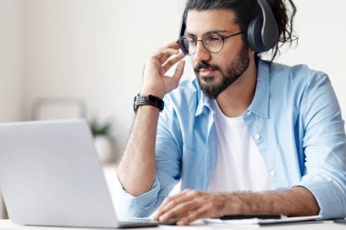 Mesafe öğrenme. Kulaklıklı genç bir adam evde dizüstü bilgisayarla ders çalışıyor, internetten giyim ya da eğitim dersleri alıyor, masada oturuyor ve bilgisayar ekranına bakıyor, boş alana bakıyor.
