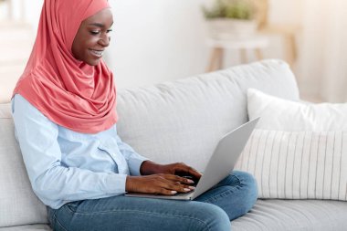Milenyum Siyahi Müslüman kadın evde dizüstü bilgisayarla çalışıyor, serbest çalışmanın keyfini çıkarıyor, dindar genç Afrikalı kadın bilgisayarla koltukta oturuyor, klavyeye yazı yazıyor, yan görünüm