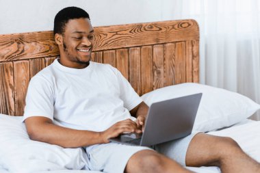 Mutlu siyah serbest çalışan adam dizüstü bilgisayarda çalışıyor internetten interneti tarıyor evdeki yatak odasında oturuyor. Uzak Bilgisayar İşi ve Serbest Kariyer Konsepti