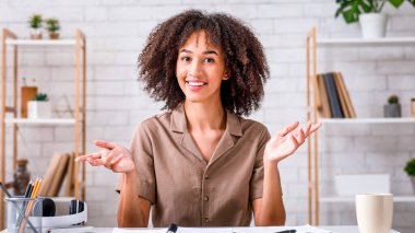 Giyin, ders ver, blog yaz ve ders al. Mutlu Afro-Amerikalı kadın el kol hareketi yapıyor ve kameraya bakıyor, salgın sırasında oturma odasında masada fincan, not defteri ve kalemle oturuyor, fotokopi çekiyor.