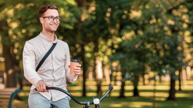 Şehre modern bir yolculuk. Gülümseyen adam elinde kahve bardaklarıyla parkta bisikletle geziyor sonbaharda, panorama, kopyalama alanı