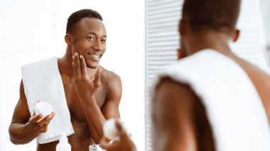 Mutlu Siyahi Adam Yüzü Nemlendirici Kullanıyor Kozmetik Krem Kavanozu, Yüzü İçin Bakıyor Evdeki Modern Banyo 'da Üstsüz Ayna Yapıyor. Erkek derisi ve güzellik rutini. Panorama