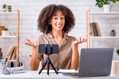 Covid-19 salgını sırasında modern blog yazarlığı ve hobisi. Neşeli Afro-Amerikan kadın takipçiler için video kaydetti ve oturma odasının iç kısmında dizüstü bilgisayarlı akıllı telefonun webcam görüntülerine baktı.