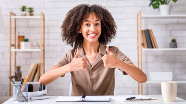 Ofiste iyi iş çıkardın. Mutlu Afro-Amerikan kadın oturma odasının iç kısımlarında kameraya başparmak gösteriyor.