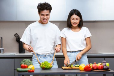 Neşeli Koreli çift taze sebze salatası yapıyor. Sağlıklı yemek hazırlıyorlardı. Modern mutfaktaki sebzelerin yanında duruyorlardı. Aile Yemekleri ve Beslenme Tarifleri