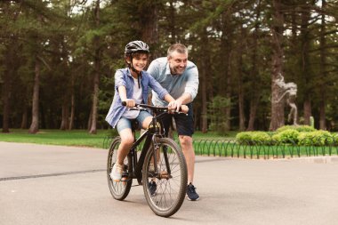 Boş zaman kavramı. Neşeli yetişkin baba portresi oğluna parkta bisiklet sürmeyi öğretiyor, sevgi dolu aile hafta sonları birlikte vakit geçiriyor, yazın dışarıda eğleniyor.