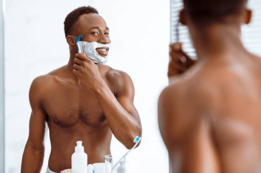 Tıraş köpüğüyle kaplı gömleksiz Afrikalı adam tıraş köpüğüyle kaplı. Sabahları evdeki banyoda aynanın yanında jilet kullanıyor. Erkek Cilt Bakımı Güzellik Programı. Seçici Odaklanma