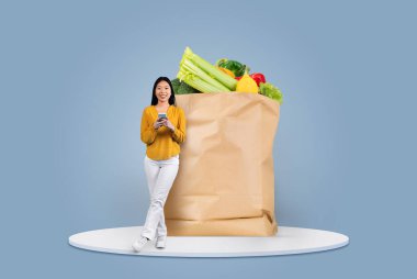 Mutlu bir müşteri akıllı telefon kullanırken taze sebze ve meyve dolu büyük bir kesekağıdının yanında durur. Mevcut yaşam tarzları arasında modern alışveriş alışkanlıklarını yansıtıyor.