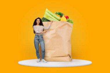 Taze sebze ve meyve dolu büyük bir kesekağıdıyla bir müşteri ayakta duruyor. Arkaplan parlak sarı, özellikle evde yapılan sağlıklı market alışverişine vurgu yapıyor..