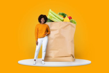Günlük kıyafet giymiş bir müşteri, sebze ve meyvelerle dolu, modern market alışveriş eğilimlerini ve ev teslimat uygulamalarını temsil eden büyük bir kese kağıdının yanında duruyor..