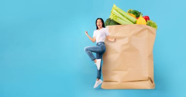 Neşeli bir müşteri, içi taze yiyeceklerle dolu büyük bir kese kağıdının yanında duruyor. Parlak mavi arka plan neşe dolu alışveriş deneyimini artırıyor, market alışverişinin yeni normalliğini yansıtıyor..