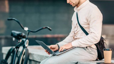 Genç bir adam bisikletinin yanında bir duvarda oturuyor, hareketli bir şehir ortamında dizüstü bilgisayarında yazı yazıyor. Yakınlarda bir kahveyle modern bir yaşam tarzını somutlaştırıyor..