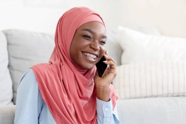 Evde dinlenirken cep telefonuyla konuşan tesettürlü, neşeli siyahi kadın, hoş bir telefon görüşmesi yapan, dindar Afrikalı kadın oturma odasında dinleniyor, hafta sonu eğlencesine çıkıyor.