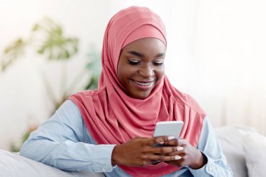 Rahat, siyah Müslüman bir kadın koltukta oturuyor ve akıllı telefonuna mesaj atıyor, rahat bir oturma odasında cep telefonuyla dinleniyor, arkadaşlarına mesaj atıyor, cep telefonu iletişiminin keyfini çıkartıyor, boş alanların tadını çıkarıyor.