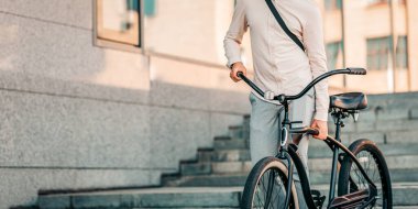 Günlük giysiler içinde genç bir adam elinde bisikletiyle şehir caddesinde yürüyor. Güneş, onu çevreleyen modern mimariyi aydınlatan sıcak bir parıltı saçar.