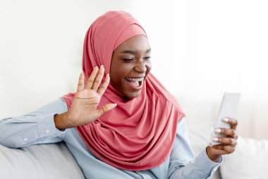 Uzaktan iletişim. Neşeli siyahi kadın evdeki cep telefonuyla video görüşmesi yapıyor, akıllı telefon kamerasına el sallıyor, arkadaşını selamlıyor, oturma odasında koltukta oturuyor, boş bir alanda oturuyor.