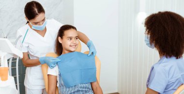 Ağız boşluğu, ortodonti ve pediatrik dişçi muayenesi ve tedavisi için hazırlık. Hemşire peçeteyi kızın üzerine koyar, Afrikalı Amerikalı doktor klinikte, panoramada, fotokopi odasında küçük kızla konuşur.