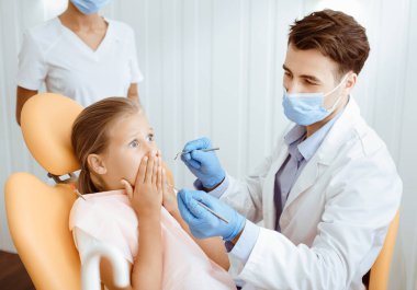 Çocuk diş hekimi ve modern klinikte bebeklerle çalışıyor. Korkmuş küçük kız hasta ağzını kapatıyor ve beyaz önlüklü, koruyucu maskeli ve lastik eldivenli dişlerin tedavi edilmesine izin vermiyor.