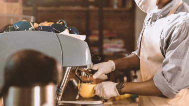 Kapatma sırasında yeni normal ve müşteri hizmetleri. Bin yıllık Afro-Amerikan önlüklü, koruyucu maskeli ve eldivenli adam, kahve makinesinde sarı kahve yapıyor.