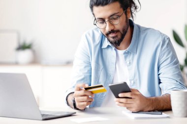 Arap serbest çalışan adam evde akıllı telefon ve kredi kartı kullanıyor, faturaları internet üzerinden ödüyor, genç doğulu adam dizüstü bilgisayarla masada oturuyor, para akışı ve alımlar için mobil uygulama kullanıyor, ücretsiz alan kullanıyor.