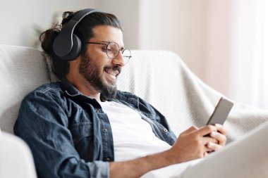 Ev Tatili. Kablosuz kulaklıklı, akıllı telefonlu genç adam kanepede dinleniyor, müzik dinliyor ve arkadaşlarıyla mesajlaşıyor, ev içi eğlencenin tadını çıkarıyor, rahat bir koltukta dinleniyor, boş bir alanda dinleniyor.
