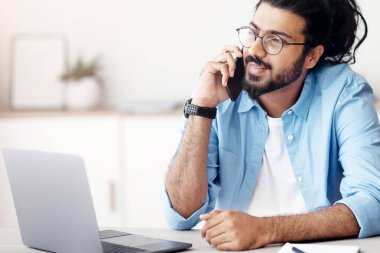 Milenyum Hintli adam cep telefonuyla konuşuyor ve merkez ofiste dizüstü bilgisayar kullanıyor, Batılı serbest çalışan adam müşterisiyle cep telefonu görüşmesi yapıyor, projeyi tartışıyor, masada oturuyor, boşluğu kopyalıyor.