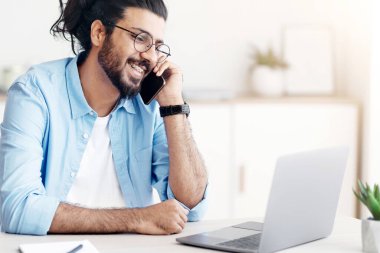 Uzak bir kariyer. Gülümseyen Doğulu serbest çalışan adam cep telefonuyla konuşuyor ve merkez ofiste dizüstü bilgisayarla çalışıyor, uzaktan çalışma fırsatlarının tadını çıkarıyor, masada oturuyor, bilgisayar ekranına bakıyor, boş alana bakıyor.
