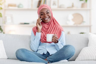 Hafta sonu eğlencesi. Neşeli siyah Müslüman kadın cep telefonuyla konuşuyor ve evde kahve içiyor, rahat koltukta dinleniyor, genç Afrikalı İslamcı kadın oturma odasında dinleniyor,