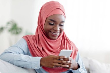 Mobil Teklif. Neşeli siyahi Müslüman kadın evde akıllı telefondan mesajlaşıyor, arkadaşlarıyla mesajlaşıyor, oturma odasında rahat bir koltukta dinleniyor, yeni uygulamalara bakıyor, kablosuz internet kullanıyor, yakın çekim yapıyor.