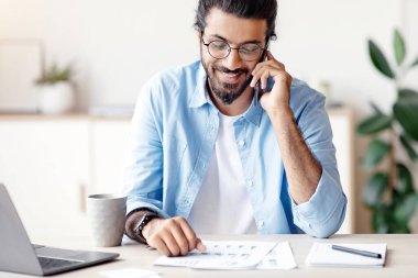 Hindistanlı serbest çalışan, kağıtları kontrol eden ve ofisteki bilgisayarda çalışırken cep telefonuyla konuşan müşteriyle konuşan, masasında oturan, gülümseyen, Plesant 'lı telefonları olan, bedava uzay boşluğu olan bir adam.