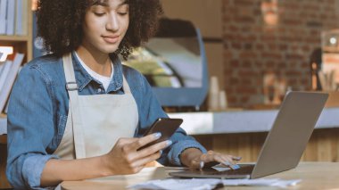 Kahve işinin sahibi sosyal medyayla muhasebe bölümünde çalışıyor. Önlüklü Afro-Amerikalı ciddi kadın akıllı telefona bakıyor ve modern kahve dükkanı Panorama 'nın iç kısmında dizüstü bilgisayar kullanıyor.