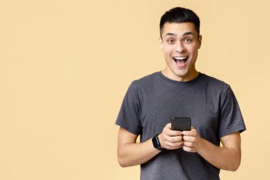 Başarı duyguları, zafer, nakit para ve kazanmakla ilgili harika bir mesaj. Kahverengi tişörtlü mutlu genç çocuk sevinçle bağırıyor ve elinde akıllı telefon tutuyor, bej arka planda izole edilmiş, stüdyo fotoğrafı.