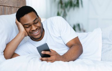 Cep telefonu kullanan gülümseyen Afrikalı adam interneti tarıyor ve telefonda mesaj yazıyor evde yatıyordu. Sosyal medyada akıllı telefon ağı olan siyah adam. Yeni Mobil Uygulama Ters Konsepti