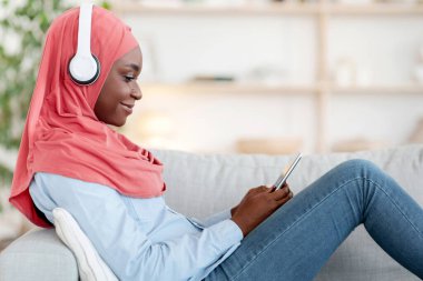 Boş Zaman Teknolojileri. Kablosuz kulaklıklı siyahi kadın evde akıllı telefonuyla dinleniyor, müzik dinliyor ve internette geziniyor oturma odasında rahat bir kanepede dinleniyor.