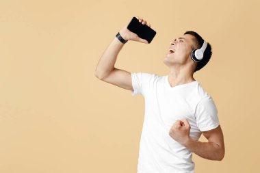 Kablosuz kulaklıklı havalı genç adam karaoke söylüyor, akıllı telefonu mikrofon olarak kullanıyor, bej arka plan, kopyalama alanı