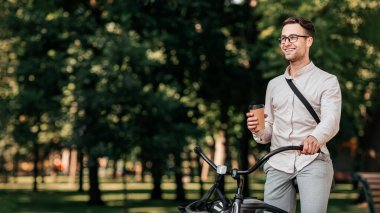 Genç bir adam yeşil şehir parkında sakin bir gezinti yaparken elinde kahve fincanı tutarak tesadüfen bisikletine yaslanıyor. Güneş ışığı ağaçların arasından süzülür, canlı bir atmosfer yaratır, uzayı taklit eder.
