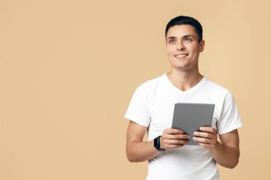 Modern teknoloji yönetimi, dijital cihazlar ve sosyal ağlar. Akıllı saati olan arkadaş canlısı genç blogcu adam tablet kullanıyor ve boş alana bakıyor, açık kumlu arka planda izole edilmiş.