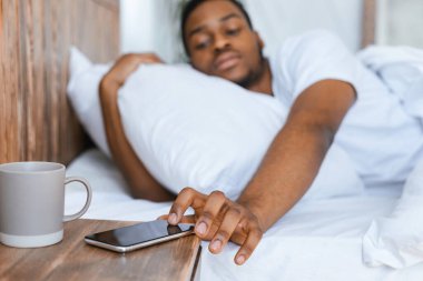 Uykucu siyah adam akıllı telefonu kapatmaya çalışıyor. Alarmı kapatıyor. Sabah erkenden uyanıyor ve sabahın köründe yatak odasında yatıyor. Uyku eksikliği ve dinlenme kavramı. Seçici Odaklanma, Sığ Derinlik