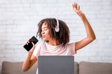 Eğlenceli müzik molası ve vokal uygulaması. Güzel Afro-Afrikalı kulaklıklı, gözleri kapalı bir Amerikalı kadın akıllı telefonda boş ekranla şarkı söylüyor, oturma odasında dizüstü bilgisayarlı bir masada oturuyor.