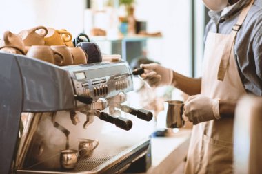 Modern kafede kahve ekipmanlarıyla çalışıyorum. Önlüklü ve koruyucu maskeli Afrikalı Amerikalı kahveci, sabahları modern kafe içinde kahve makinesiyle kahve yapıyor.