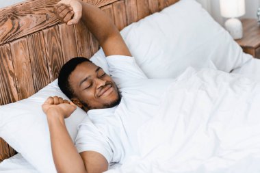 İyi sabahlar. Dinlenmis siyah adam ellerini uzatarak uyaniyor rahat bir yatakta uzaniyor, gözleri kapali bir sekilde gülüyor. İyi bir uykudan ve dinlenmeden sonra iyi uyuyan adam uyanıyor. Yüksek Açı Görünümü