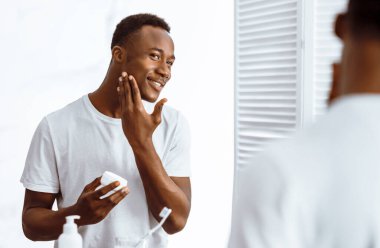 Yakışıklı siyah adam yüz kremi kullanıyor ve cildini nemlendiriyor. Sabahları banyoda aynanın yanında dikiliyor. Erkek hijyeni, Seflcare ve cilt bakımı konsepti. Seçici Odaklanma