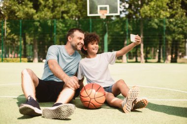Mutlu baba-oğul portresi. Akıllı telefon kullanarak selfie çekiyor. Spor sahasında oturuyor.