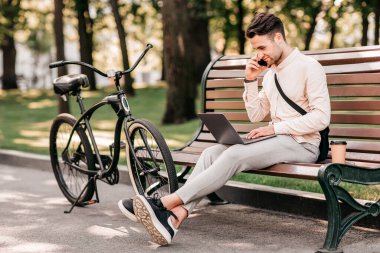 Müşteriyle önemli bir konuşma. Gündelik kıyafetleri olan yakışıklı genç adam dizüstü bilgisayarda çalışıyor ve telefonla konuşuyor. Bisikletin yanında, açık havada, yan gözle, boş alanda otururken.