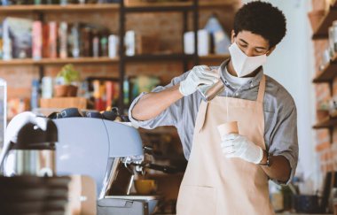 Modern kafe, küçük işletmeler ve sosyal mesafeler. Milenyum Afrikalı Amerikalı önlüklü, koruyucu maskeli ve beyaz eldivenli barmen, kafenin içindeki kağıt bardağa süt döküyor.