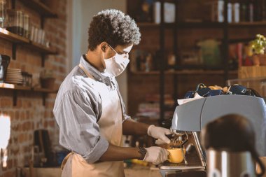 Covid-19 sırasında yeni müşteri hizmetleri kuralları, güvenli olun. Konsantre olmuş Afro-Amerikan erkek önlüklü, maske kahve makinesinin üstünde latte yapıyor kafenin arka planında, güneş parlaması