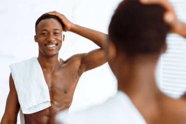 Gömleksiz Afrikalı Amerikalı adam, duş aldıktan sonra aynaya bakıp modern banyoda yansımasına gülümsüyor. Erkek Güzelliği ve Saç Bakımı rutini. Seçici Odaklanma